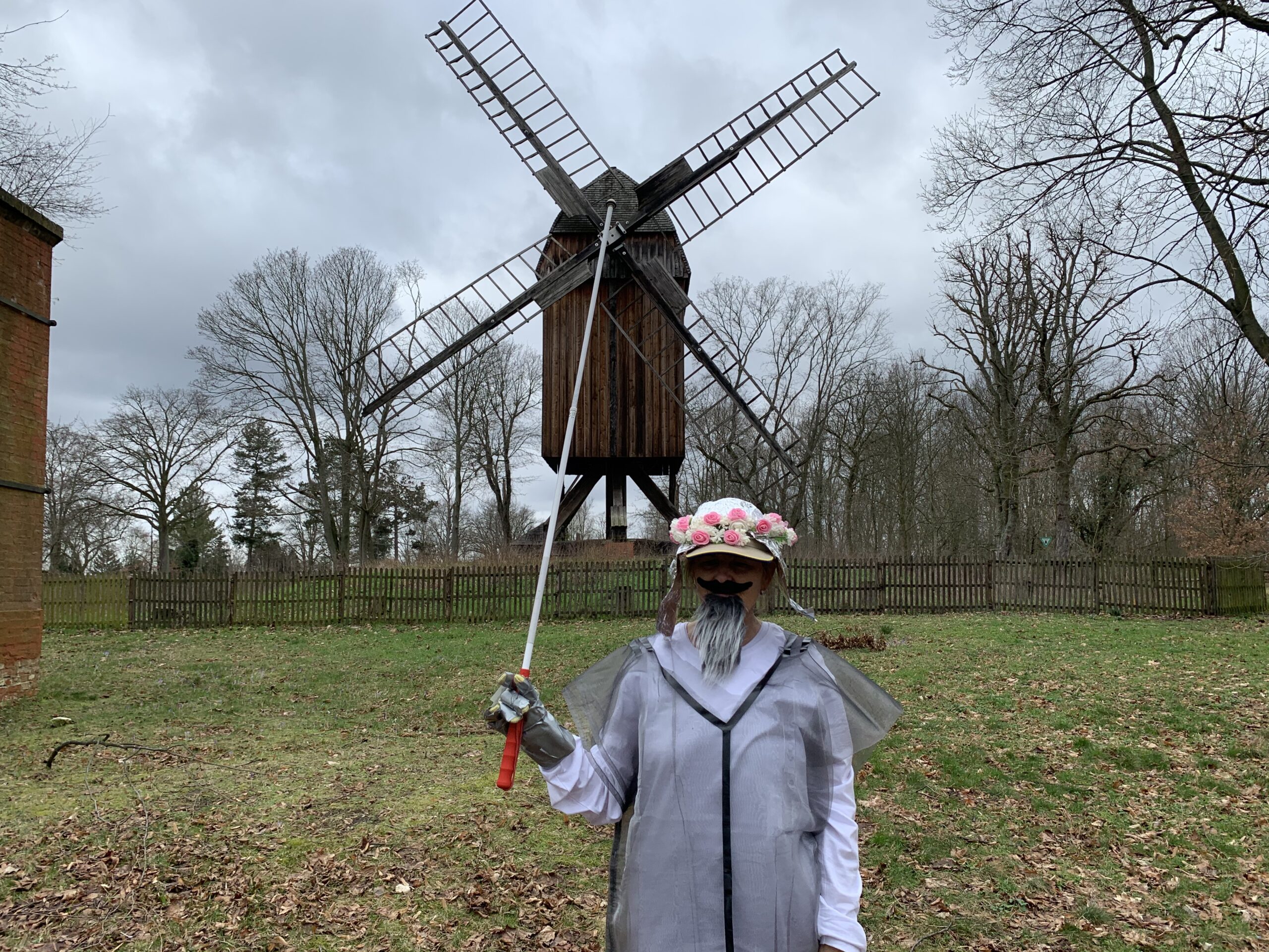 Don Quijote vor einer Windmühle. Eine Person in einem grau-durchsichtigen Regencape auf weißem Hemd mit grauem Bart und einer weißen, blumenumkränzten Mütze hält einen Stab in einem Metallhandschuh senkrecht nach oben, sodass dieser optisch die Mitte der vier Windräder von der Windmühle hinter ihr trifft.