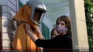 ZOOM Screenshot: Lavinia fühlt den orangenen Schutzanzug an der Halsgegend und schaut in die Kamera. Sie steht auf der Veranda und der Anzug mit dem weißen Helm hängt an der Veranda-Wand.