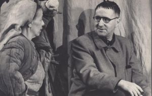 © Hainer Hill © AdK, Berlin. Berliner Ensemble "Mutter Courage und ihre Kinder", 1951. Helene Weigel lehnt sich mit einem hocherhobenen Arm an die Kulisse und schaut mit der anderen Hand an der Hüfte zu Bertolt Brecht. Dieser schaut sie von der Seite an. Er hat kurze dunkle Haare, eine schwarz-umrandetet Brille, trägt ein weites Arbeiter-Sakko und hält eine Zigarre in seiner rechten Hand.